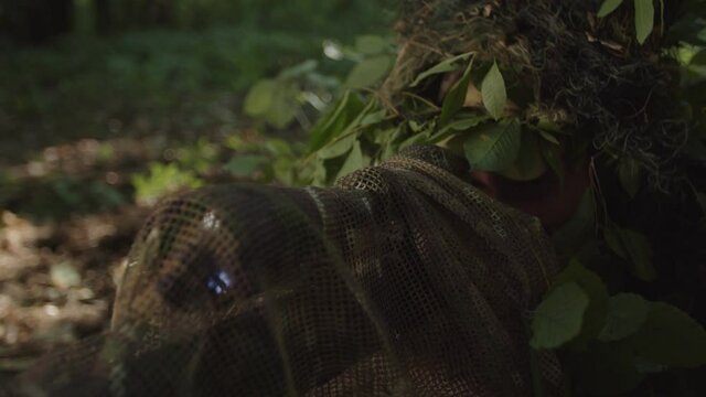 Young military marksman in disguise sniper coat lying on equipped observation position, using camouflaged sniper rifle and looking through scope of optical sight, taking aim in forest at daybreak