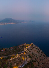 Large panorama. Wide aerial view of Alanya bay, Turkey, Summer sunset evening, night time. Travel and vacation. Kalesi Castle. Ships and boats at sea. City lights. Streets and houses.