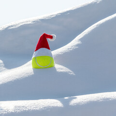 Christmas tennis ball in red Santa Claus hat on snowy hills on sunny winter day. New Year and...