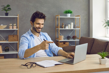Happy positive handsome man sitting at table at home smiles and points index finger at screen of...