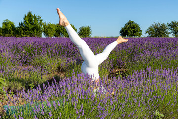 Woman in a white leotard doing yoga warm-up exercises in a blooming lavender field. Lying on the ground, she trains her legs by lifting them in the air past the lavender flowers.