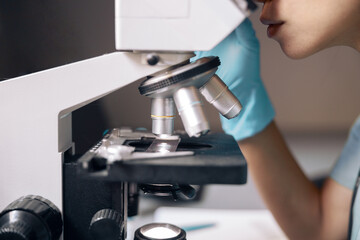 Employee in gloves looks at sample through microscope at workplace in laboratory