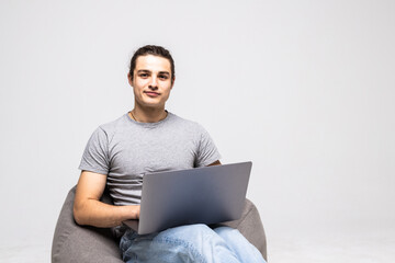 Nice photo of handsome young man using laptop and sitting on big cushioned frameless chair isolated on white background.