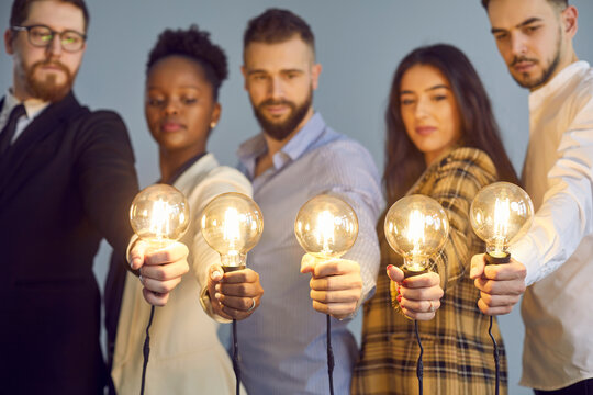 Multiethnic Team Of Serious Determined Intelligent Business People, Workers, Corporate Employees Looking At Glowing Edison Light Bulbs, Ready To Implement Good Genius Breakthrough World Changing Idea