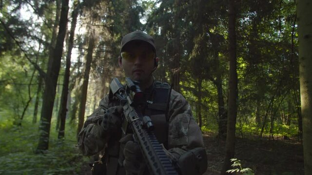 Close-up portrait of fully equipped soldier in camouflage using assault rifle, while military group of man and woman moving behind with guns ready to apply in forest at daybreak