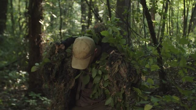 Young soldier weaving leaves into military ghillie suit cloak, wearing up tactical sniper camouflage hood cape, lying down on position, looking through scope of optical sight, taking aim in forest