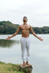 Young Brazilian man standing beside a lake, shirtless, with arms open and looking up.