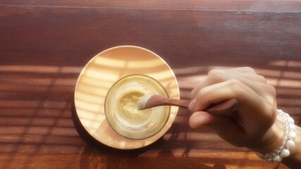 Elegant female hand stirring affogato coffee with a wooden spoon. Delicious morning caffeine drink for a woman. Avogato in the golden light through shutters on a wooden table. Lady enjoying beverage.