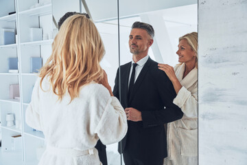 Beautiful mature woman adjusting suit of her husband while both standing in front of the mirror at...