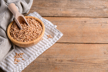 Wholegrain uncooked raw spelt farro in a bowl on napkin with wooden scoop on wooden background, top view