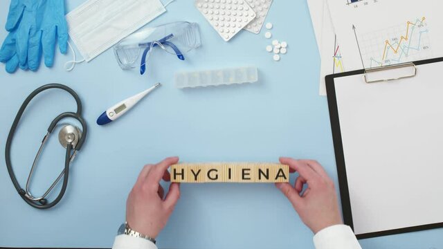 Hygiene is word written on wooden cubes that lie on table with medicines and other medical supplies. Doctor's hands take inscription, closeup. Concept of hygiene compliance