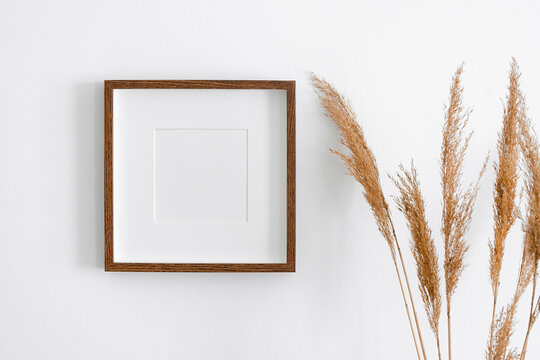Square wooden frame mockup for artwork, photo, print and painting presentation. White interior walll with dry grass decoration.