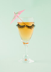 Tall  wine glass with black lashes containing a orange drink and a pink umbrella. Minimalist summer concept on a light green background