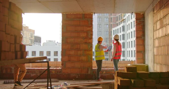 Caucasian male and female constructors in helmets and vests talking and discussing plan draft at building side. Asian male constructor started to discuss plan draft all together on tablet.