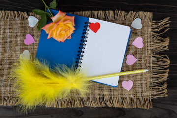 On an open notebook lies a rose, a pen with a bird's feather and confetti in the form of hearts.