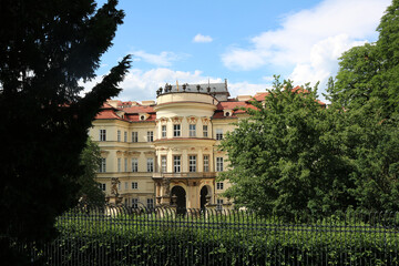 Lobkovicz Palace in Prague illuminated by the bright sun among the greenery of trees against the background of an azure sky and light clouds