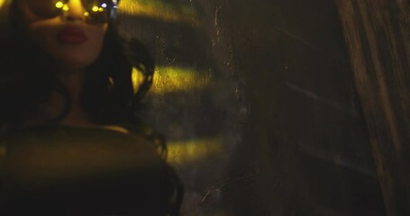 Stylish brunette girl in sunglasses dancing near the wall with jalousie lines light . Trendy young woman dancing single in club . Hot girl with long hair performs modern dance against concrete wall .