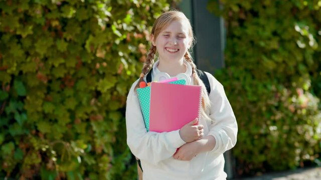 Portrait of smiling modern child in white sweatshirt with workbook and headphones near green school outdoors in the city.