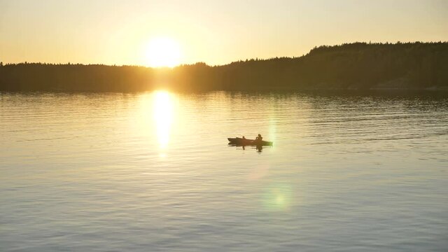 silhouette of person with little child sailing small kayak along large tranquil lake against dark forest at bright sunset