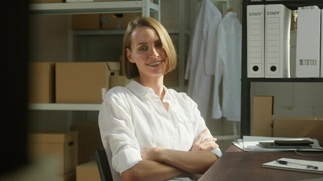 Young smiling woman sitting in storage, female employee of warehouse wearing formal office clothes, looking in camera on background of cardboard boxes. Delivering and logistics, small woman business.
