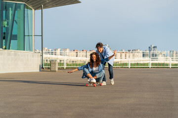 Obraz premium Young caucasian couple enjoy skating together on street. Hipster man pushing back of woman sitting on longboard. Cheerful lovers or friends have fun outdoors over city skyline. Urban lifestyle concept