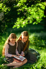 Two schoolgirls in school uniforms are reading a book while sitting on the green grass in the park. Do homework