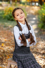 Portrait of a cute schoolgirl in a school uniform sitting on a wooden chair among the autumn...