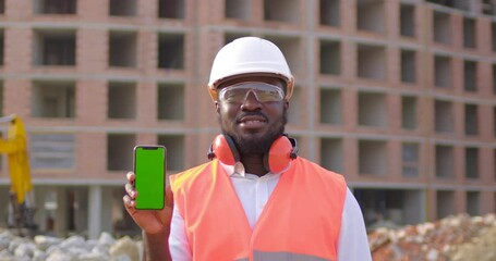 African amirican man engineer shows green phone screen, standing in a helmet, protective glasses and headphones. Engineer holds the phone with the app and looks at camera, smiling.