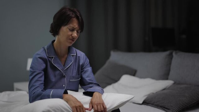 Frustrated mature woman in blue pajamas sitting on bed and hugging pillow. Tired dark-haired lady suffering from suffering from insomnia at night.
