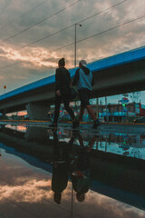 Dos personas caminando sobre un charco de agua con un puente de fondo a la luz del atardecer con presencia de nubes