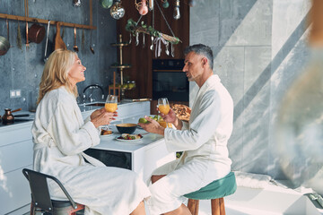 Beautiful mature couple in bathrobes enjoying breakfast together while spending time in the domestic kitchen