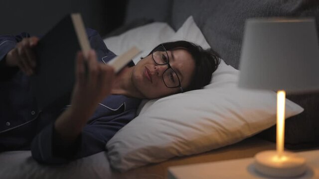 Pleasant woman in pajamas and eyeglasses reading book in bed before sleep. Concept of people, leisure time and lifestyles.