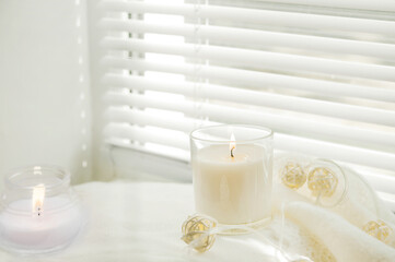 Fototapeta na wymiar Burning candles and white sweaters on the windowsill. Cozy autumn concept and a window with blinds...