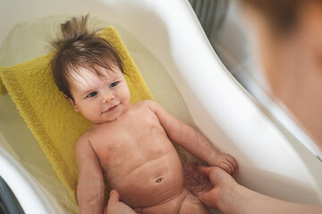 Obraz premium Small caucasian baby taking a bath at home - unknown woman mother holding her small child baby in plastic bath tub in room at home parenting and hygiene concept real people