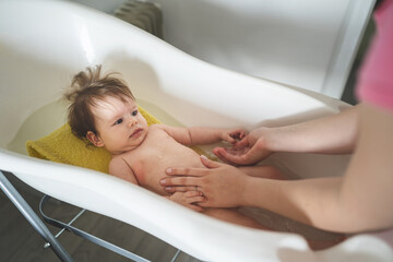 Small caucasian baby taking a bath at home - unknown woman mother holding her small child baby in plastic bath tub in room at home parenting and hygiene concept real people