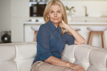 Attractive mature woman with pleasant smile looking at camera while relaxing, sitting on sofa in her modern apartment