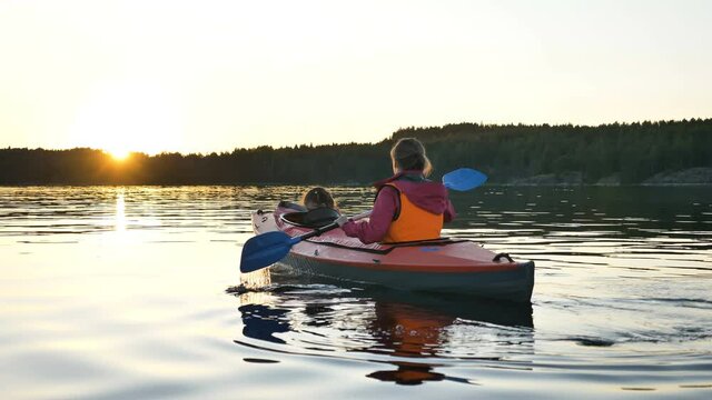 young blonde mother in orange vest rows small sports kayak with little girl along tranquil river in evening backside view