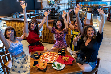 group of mature women sitting in a pub raise their hands while eating pizza