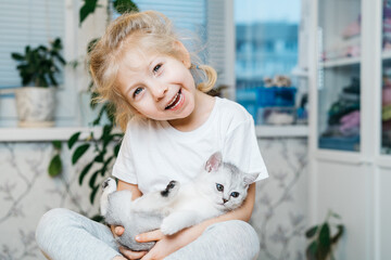 child playing with little cat. A little girl holds a white kitten. A little girl snuggles up to a cute pet and smiles while sitting in the living room of the house. Happy Children and pets.