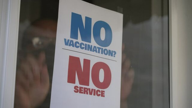 A small business owner places a NO VACCINATION NO SERVICE sign on his front door window.  	