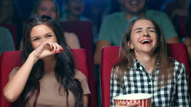 Two laughing female friend eating popcorn watching comedy film cinema rejoicing having fun