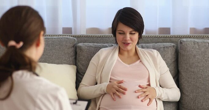 Happy beautiful young pregnant woman with big belly sitting on sofa, consulting with obstetrician gynecologist, checking up health in clinic office or consulting with doula about labor, prenatal care.