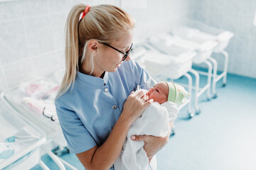 Young nurse standing in maternity ward and holding newborn baby in her arms. After birth concept.