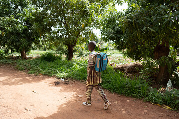 Young african boy in traditional dress and white rubber sandals on his way to school with a big...