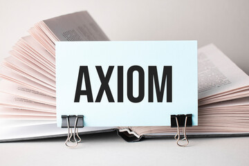 A white card with the text axiom stands on a clip for papers on the table against the background of books. Defocus