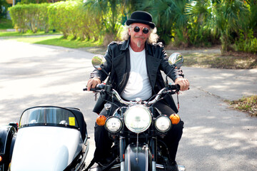 A handsome blond-haired, goatee-wearing, steampunk biker driving a black and white sidecar motorbike. Wearing leather tailcoat, bowler hat, and round sunglasses. Driving towards viewer