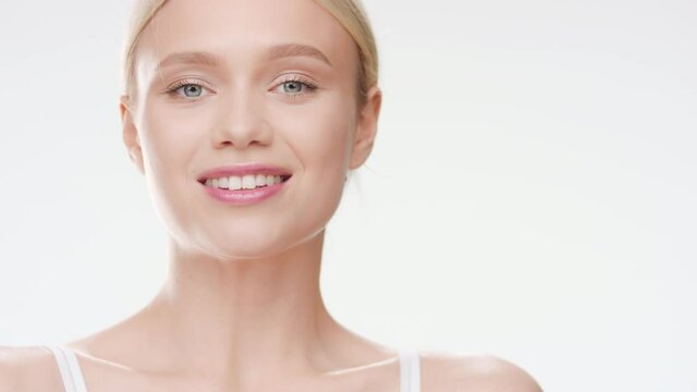 Pretty slim Caucasian young light-haired woman in white bra touches her chin turning her body and smiling wide for the camera on white background | Skincare ad concept