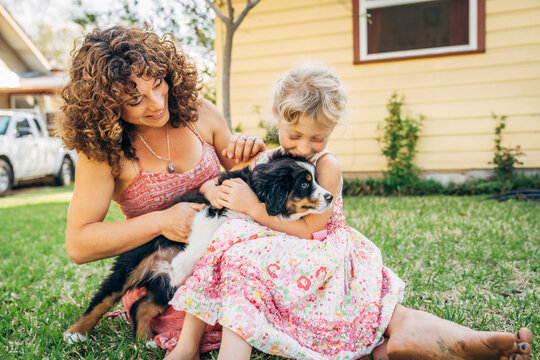 Pregnant woman and daughter sitting on grass in yard playing with puppy