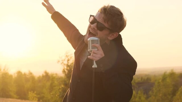 man in black glasses and black jacket sings song into shiny vintage retro microphone in nature against backdrop of sunset or dawn sunrise. rock star performs his hit with cigarette in his hand.