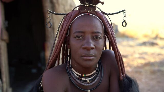 Married Himba woman looking at camera wearing traditional jewelry and Erembe headpiece at her village near Kamanjab in Namibia, Africa, slow motion shot.